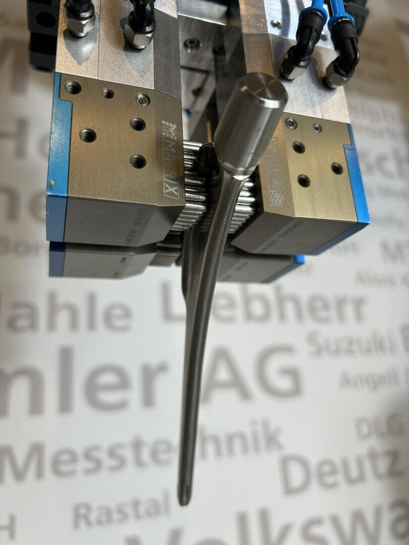 Nahaufnahme eines Robotergreifers, der ein Automobilventil aus Metall hält. Im Hintergrund ist eine verschwommene Wand mit den Namen verschiedener Automobilhersteller zu sehen, darunter Volkswagen, MAN und Deutz.