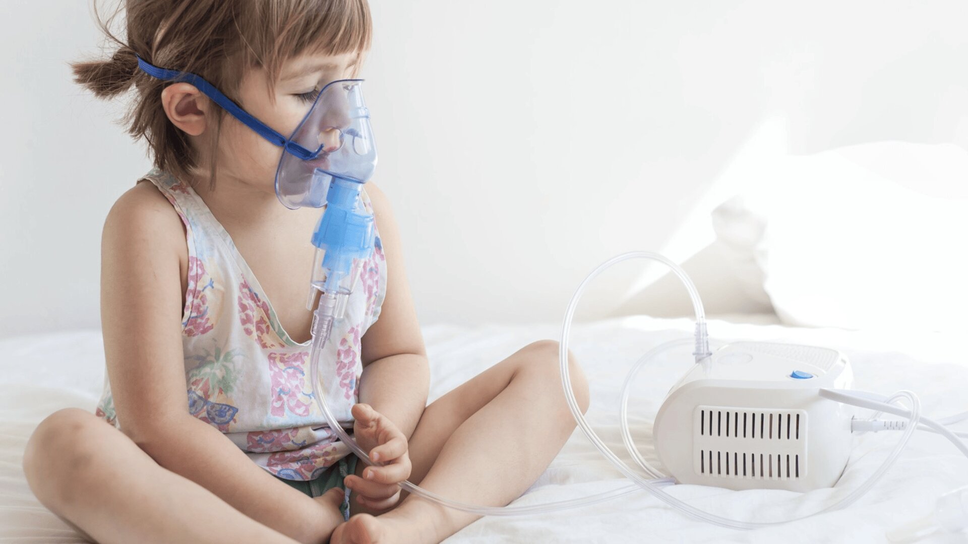 Ein kleines Kind sitzt mit einer an ein Gerät angeschlossenen Verneblermaske auf einem Bett und erhält eine Atemtherapie. Es wirkt ruhig und trägt ein geblümtes Tanktop und Shorts.