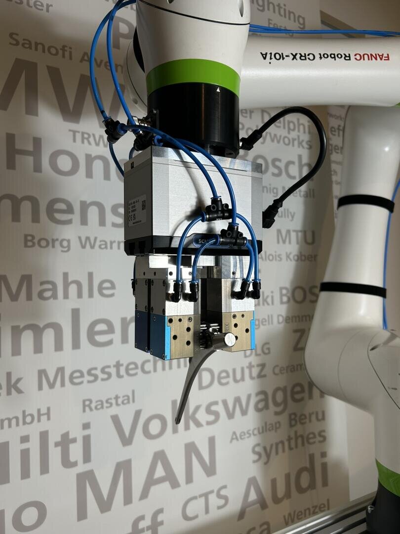 Nahaufnahme eines Roboterarms mit einem mechanischen Greifer und blauen Schläuchen, der vor einer Wand steht, an der verschiedene Namen von Automobil- und Industrieunternehmen in verschiedenen Größen und Grautönen angebracht sind.