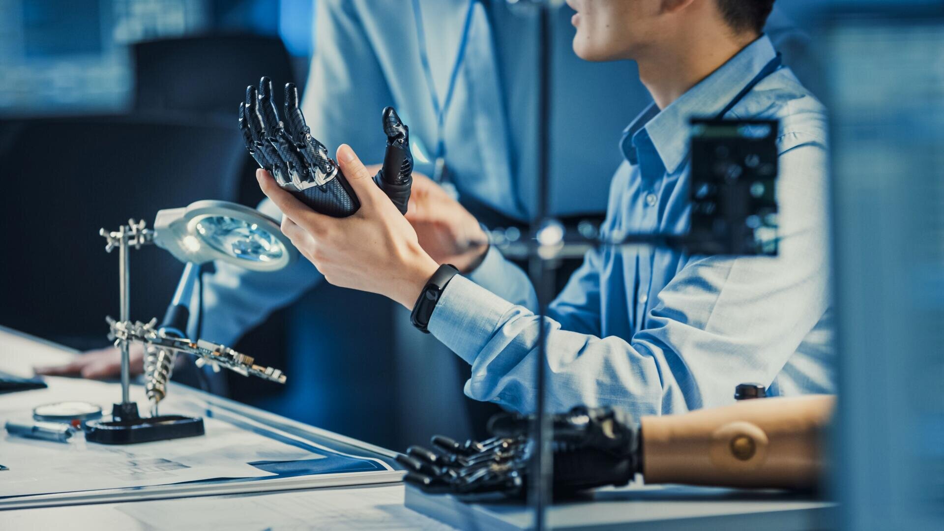 Eine Person in einem Laborkittel untersucht eine Roboterhand an einem Schreibtisch mit Werkzeugen, Plänen und einer weiteren Armprothese in der Nähe, was auf ein Robotik- oder Prothetiklabor schließen lässt.