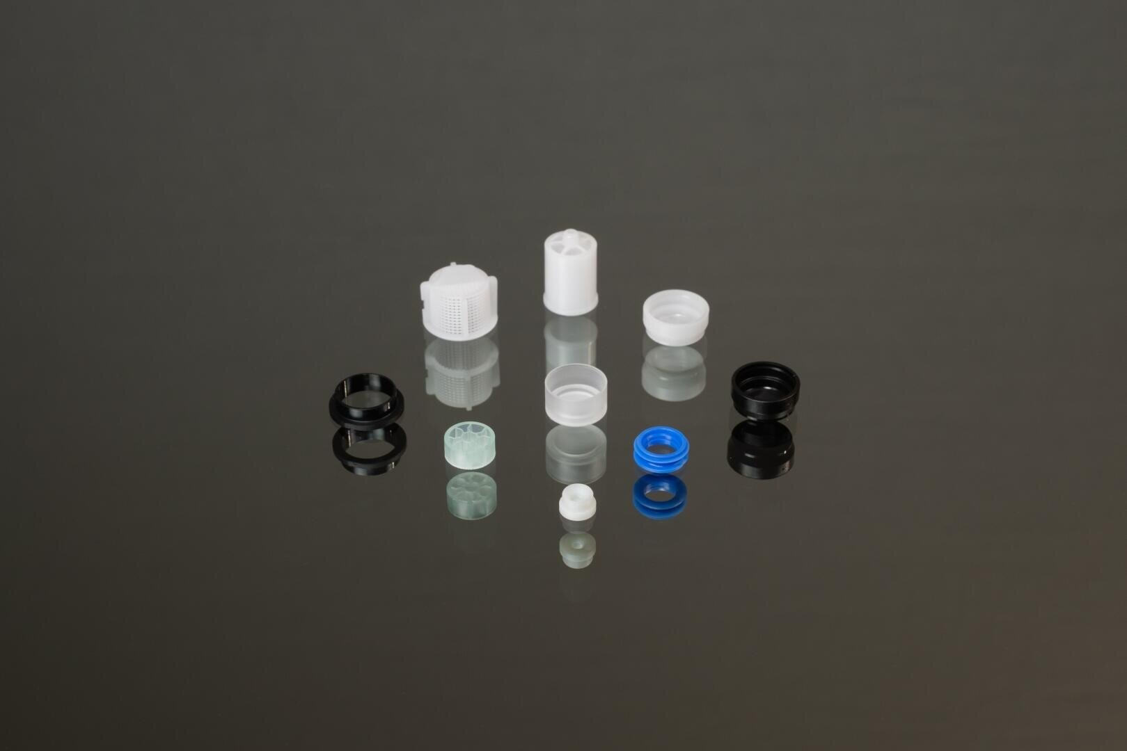 Assorted small plastic and rubber components, including rings and caps in black, white, blue, and translucent colors, arranged neatly on a reflective dark surface.
