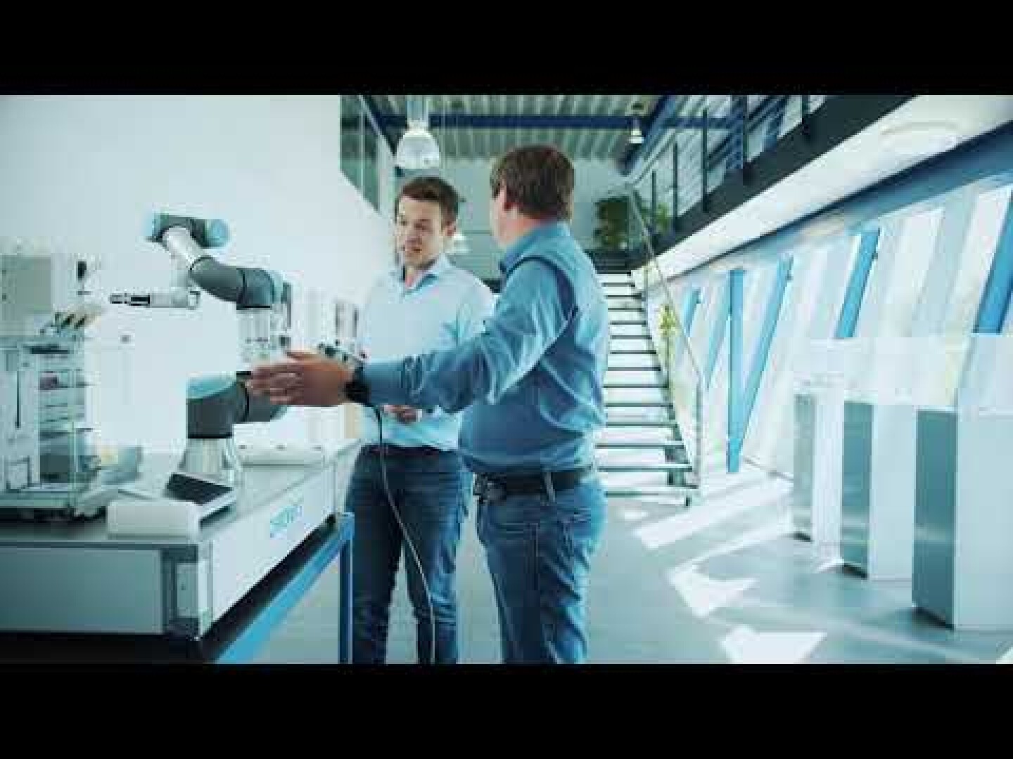 Zwei Männer in einem modernen, hellen Büro diskutieren über einen Roboterarm an einer Arbeitsstation. Einer deutet auf den Roboter, während der andere zuhört. Im Hintergrund ist eine Treppe mit Glaspaneelen zu sehen.
