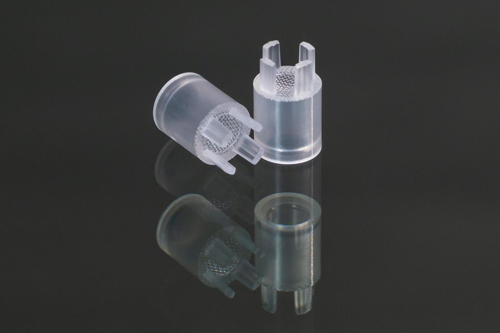 Two clear plastic cylindrical components with textured grips and open prongs are placed on a glossy black surface, creating a reflection.