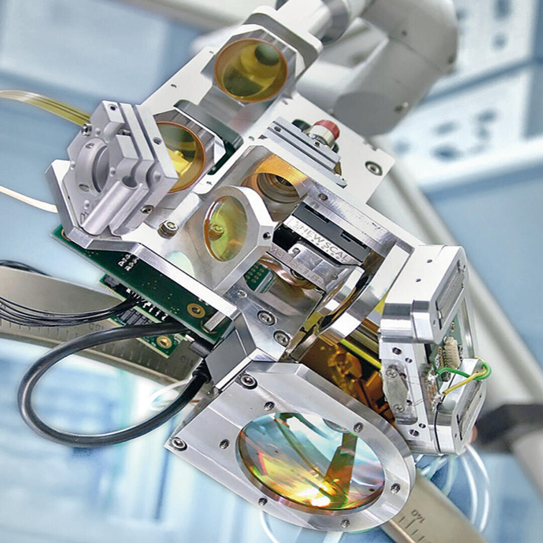 Nahaufnahme eines komplexen optischen Geräts mit mehreren Metallkomponenten, Leiterplatten und runden, bunten Glaslinsen, wahrscheinlich Teil wissenschaftlicher oder medizinischer Geräte in einer Laborumgebung.