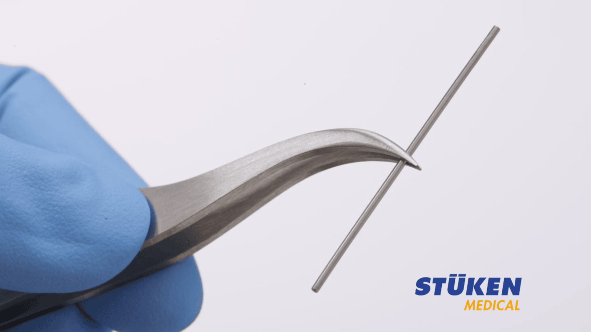 Eine behandschuhte Hand hält eine gebogene Pinzette, die einen dünnen Metallstab umklammert. In der unteren rechten Ecke ist das Logo von Stüken Medical zu sehen.