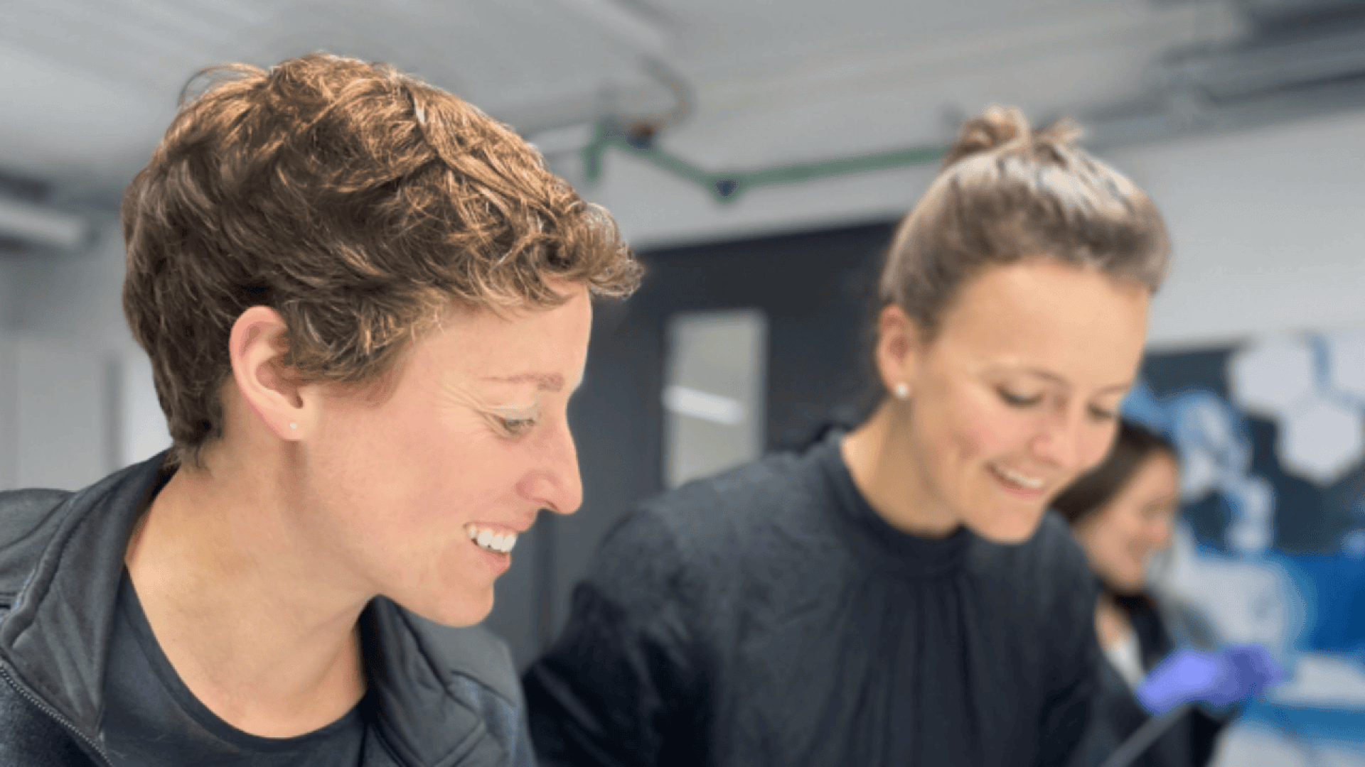 Two women smiling and looking down at something out of frame in a bright, modern indoor setting, with another person blurred in the background.