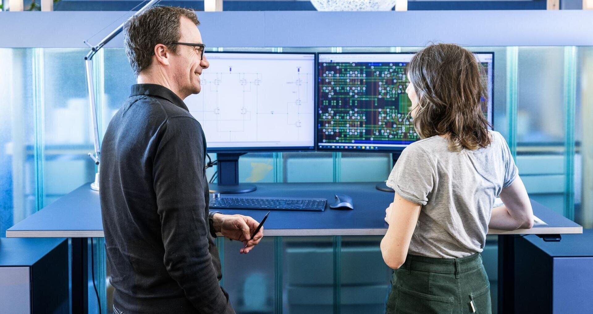 Two people are standing at a desk discussing data displayed on two large computer monitors with technical diagrams and colorful circuit diagrams in a modern office environment.