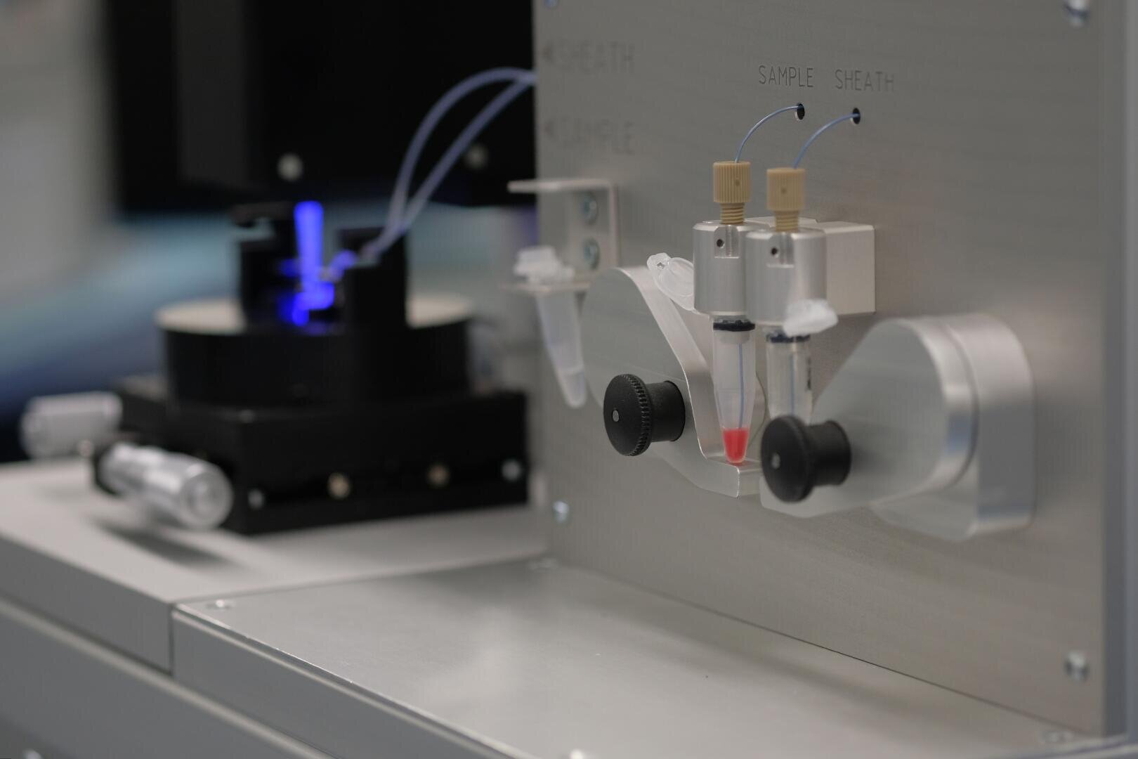 Close-up of a laboratory machine with labeled ports for sample and sheath, tubes connected to vials, and a blue light in the background, used for scientific or medical analysis.
