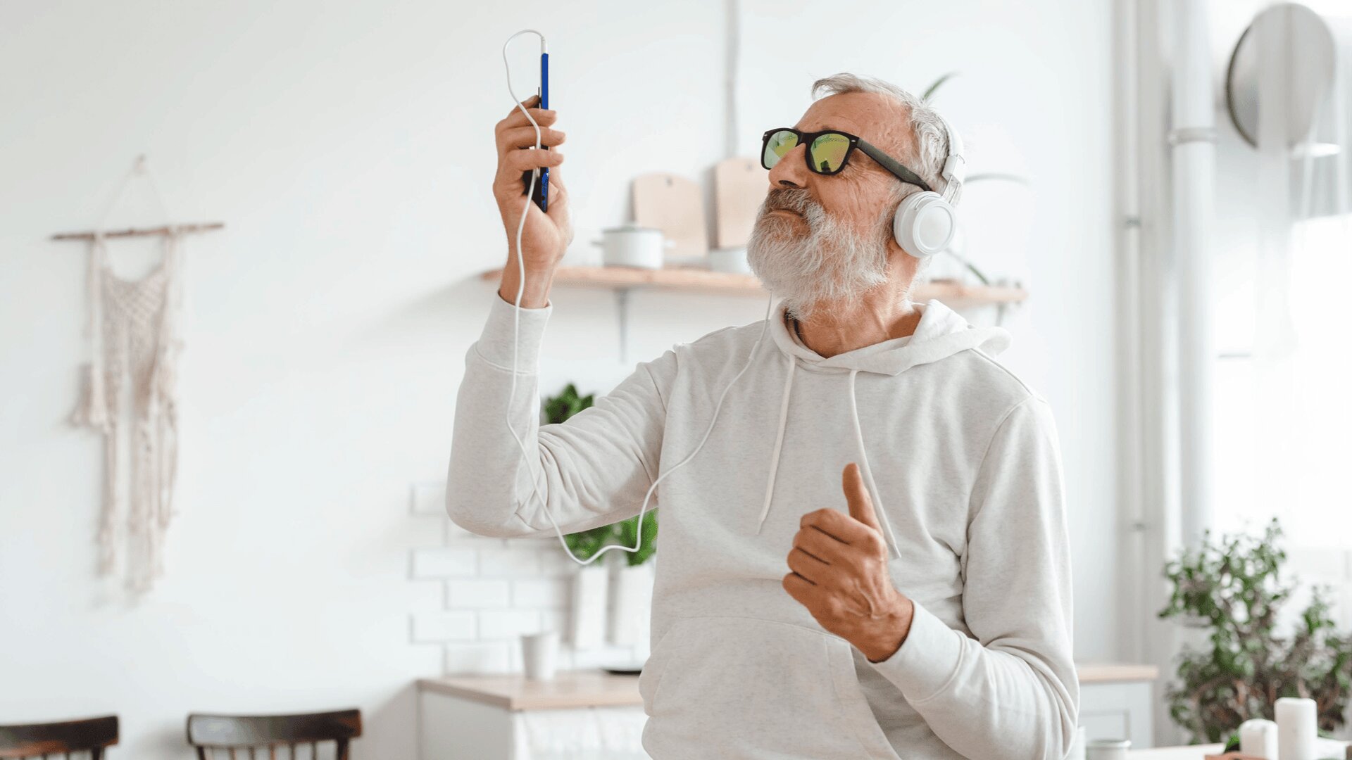 Ein älterer Mann mit Sonnenbrille und Kopfhörern, der ein Smartphone hält und Musik hört, tanzt in einer hellen, modernen Küche. Er wirkt entspannt und fröhlich, trägt einen leichten Kapuzenpulli mit Pflanzen im Hintergrund.