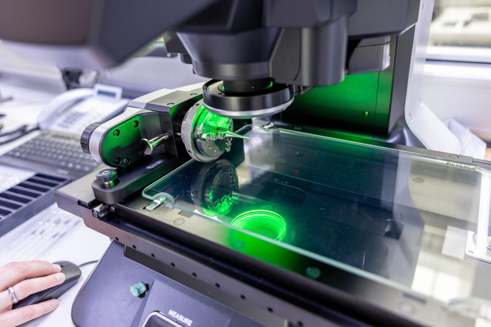 Close-up of a high-tech microscope with a slide being examined under green light in a laboratory setting, with a person's hand partially visible on the left.