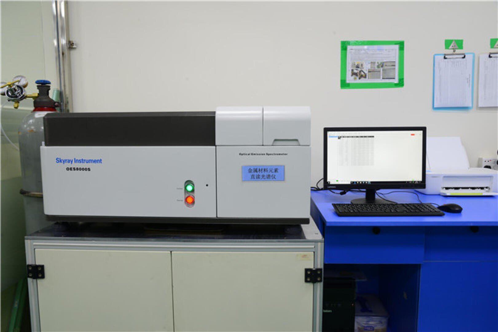 Ein Laboraufbau mit einem Skyray Instrument OES8000S-Spektrometer auf einem Schrank neben einem Computer auf einem blauen Schreibtisch, der Daten auf dem Monitor anzeigt, und Laborunterlagen an der Wand.