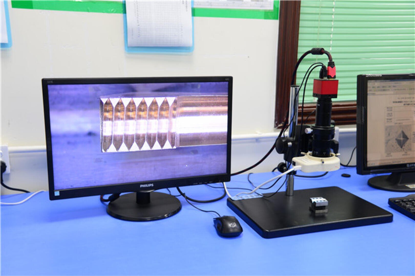 Ein Computermonitor zeigt eine vergrößerte Ansicht eines metallischen Objekts mit Rillen. Daneben befindet sich ein digitales Mikroskop über einer kleinen Metallprobe auf einem blauen Schreibtisch in einer Laborumgebung.