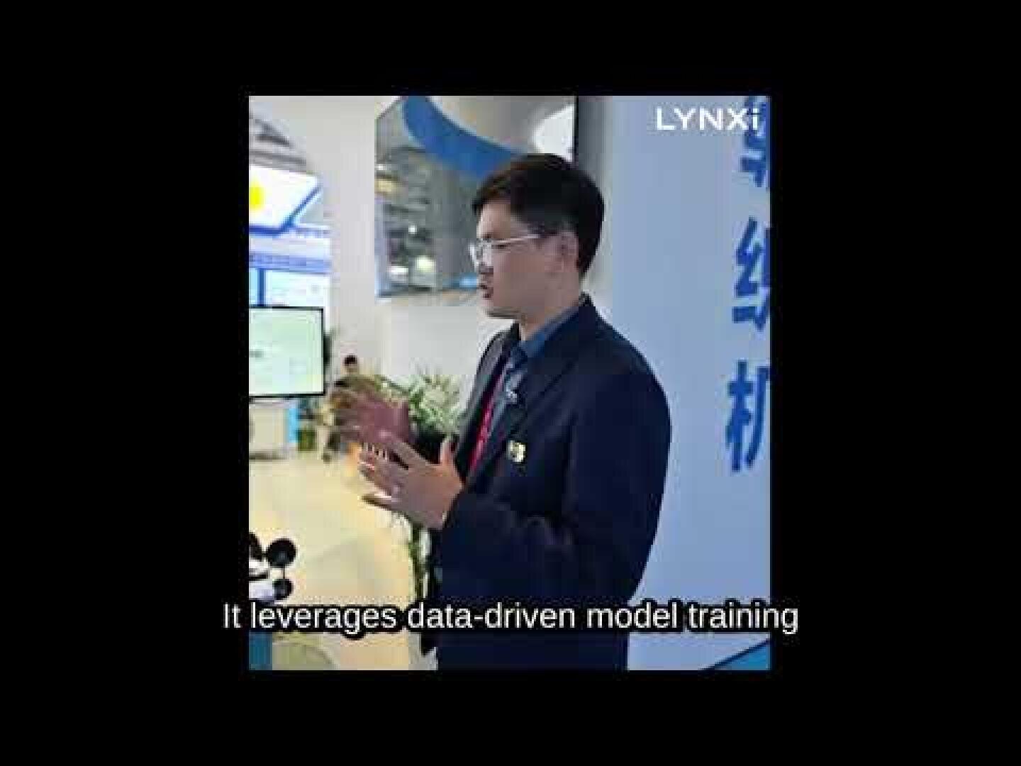 Ein Mann in einem Anzug und mit Brille spricht vor einem Display auf einer Veranstaltung oder Messe. Oben rechts ist das Wort LYNXI zu sehen, und die Untertitel lauten: "It leverages data-driven model training.