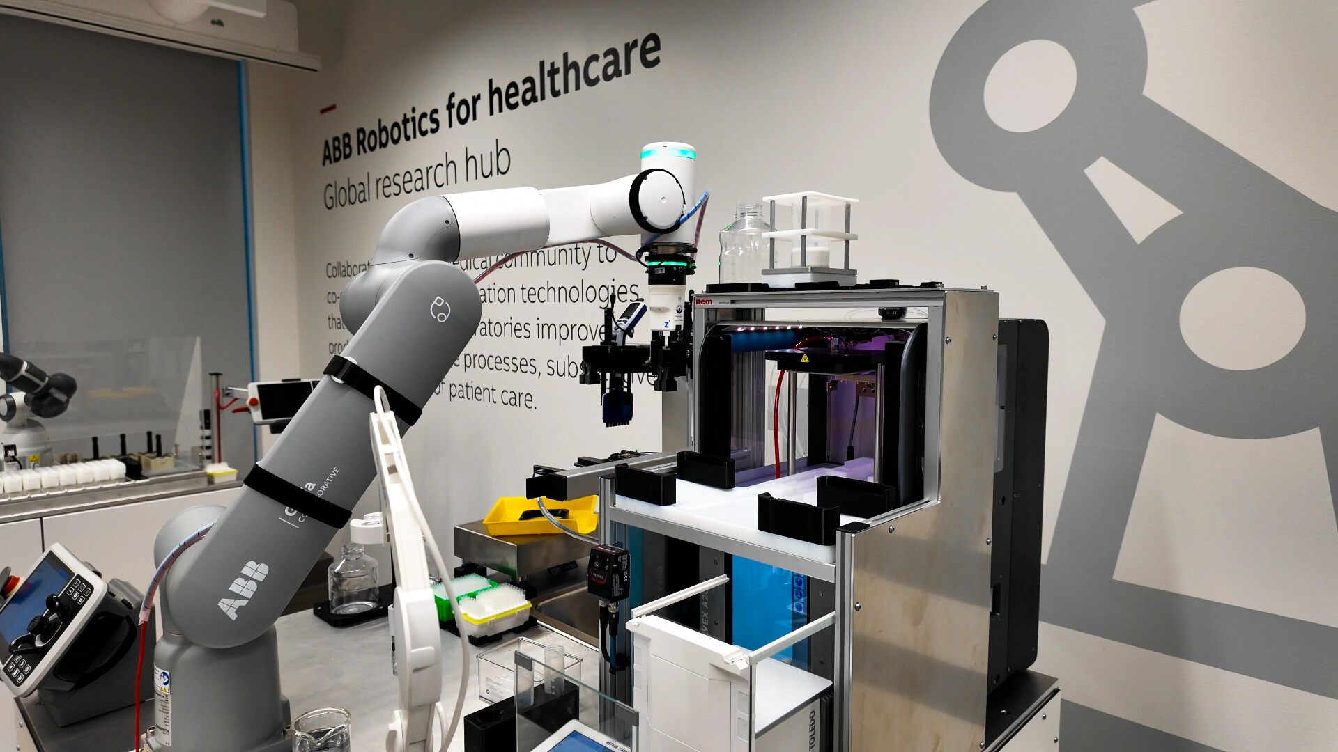 Ein Roboterarm bedient Laborgeräte in einer modernen medizinischen Forschungseinrichtung. Im Hintergrund sind an der Wand die Worte „ABB Robotics for Healthcare“ und der entsprechende Text zu sehen.