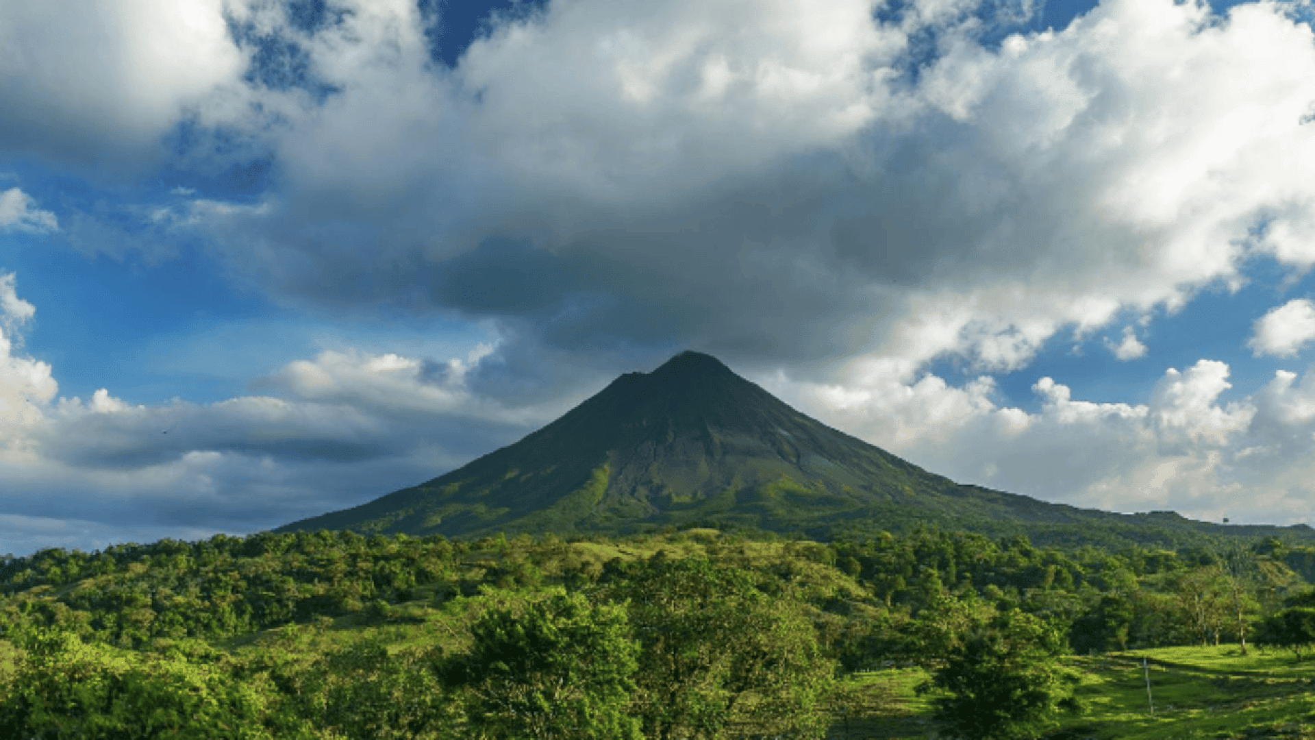 A lush green volcano rises from dense forest under a blue sky with scattered clouds, some of which cast shadows on the volcano’s slopes.
