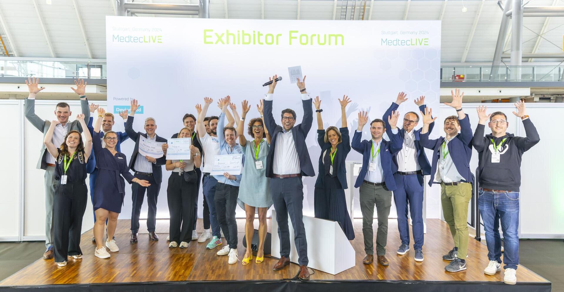 Eine Gruppe von Personen auf der Bühne des MedtecLIVE-Ausstellerforums hebt freudig die Hände zum Feiern. Einige halten Urkunden, eine Person hält ein Mikrofon. Über ihnen ist ein großes Exhibitor Forum-Schild zu sehen.