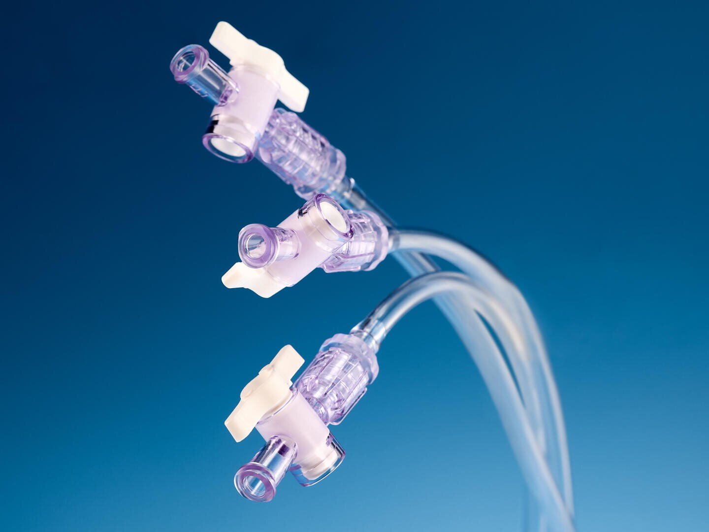 Nahaufnahme von drei klaren medizinischen IV-Hähnen mit violetten und weißen Komponenten, die an transparente Schläuche angeschlossen sind, vor einem glatten blauen Hintergrund.