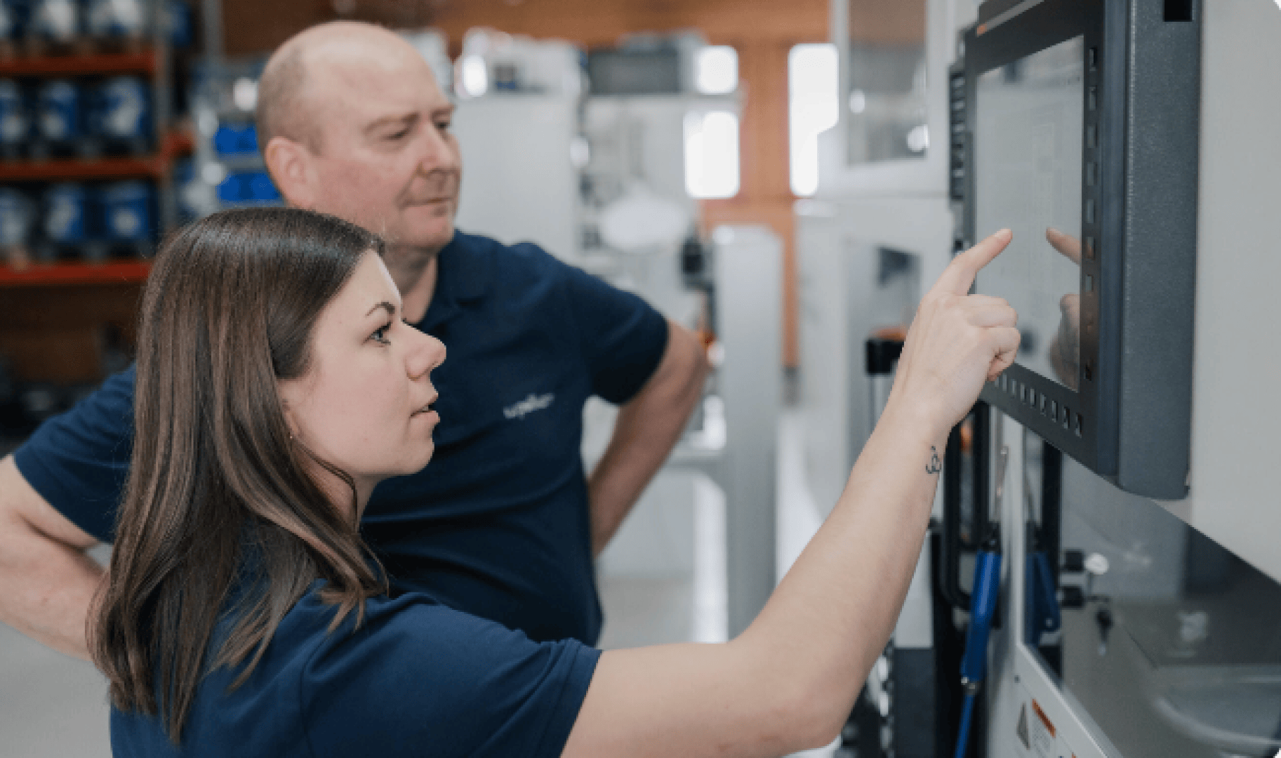 A woman points at a touchscreen on a large machine while a man stands beside her, observing. Both are wearing navy blue shirts in an industrial or workshop setting.