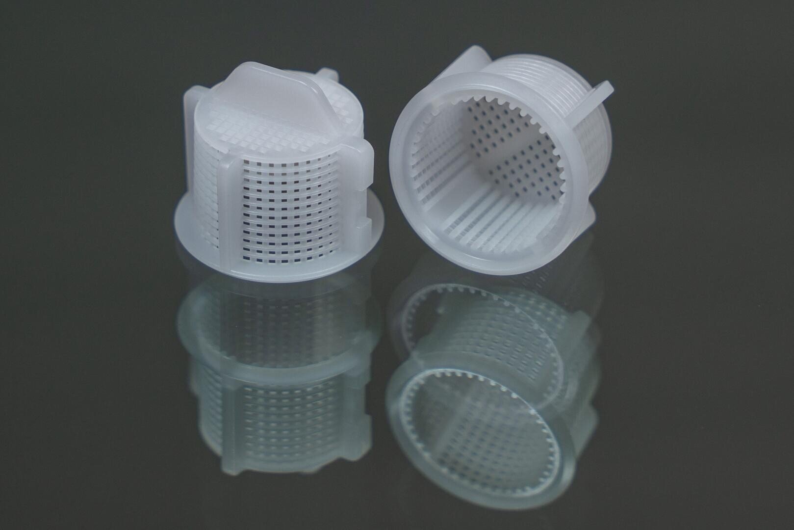 Two small, white, cylindrical plastic mesh filters with perforated sides and bases are shown on a reflective dark surface, with one filter standing upright and the other lying on its side.
