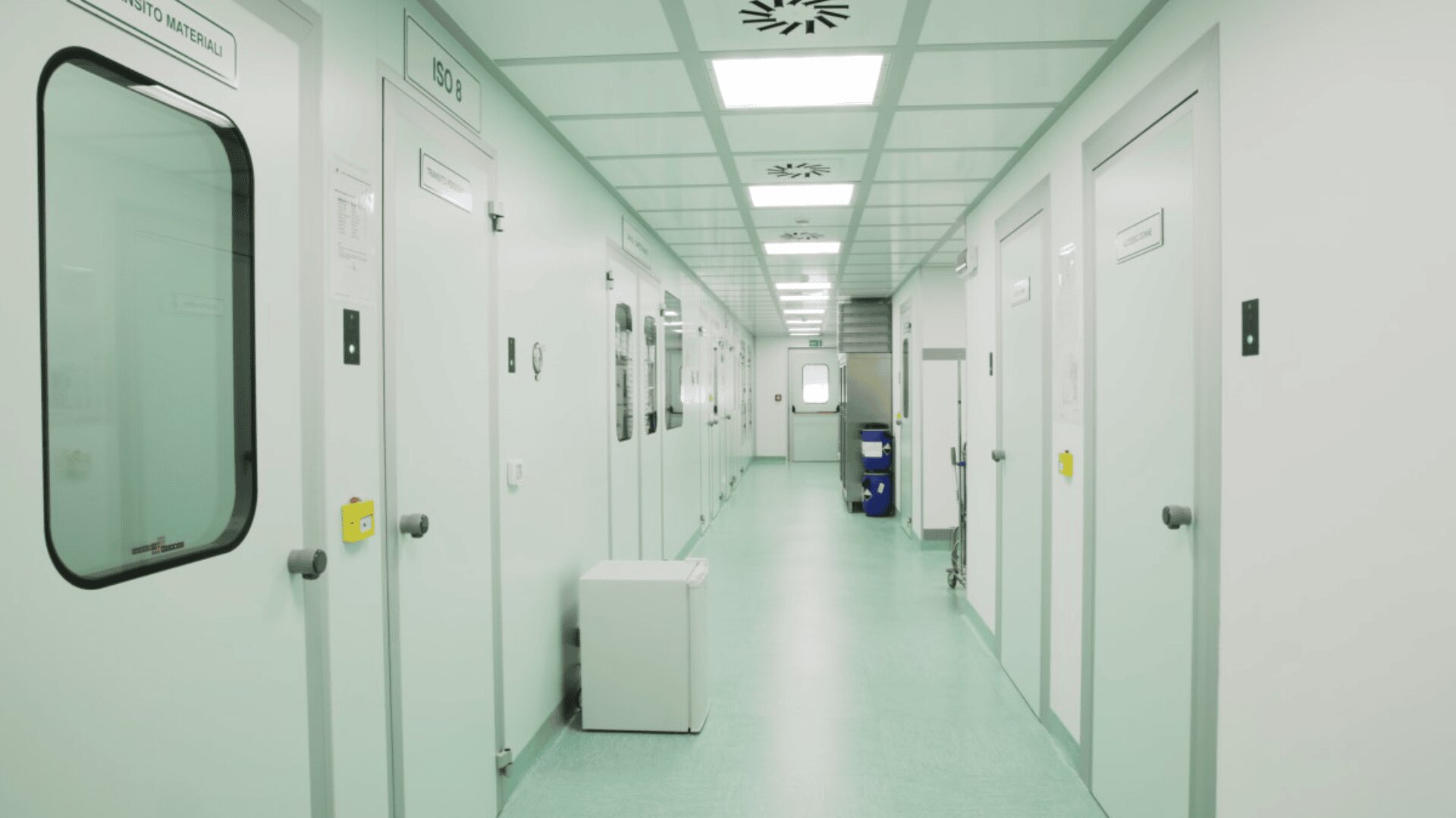 Bright, clean hospital corridor with pale green floors and white walls, lined with closed doors and medical rooms. A medical cart is visible at the far end of the hallway.