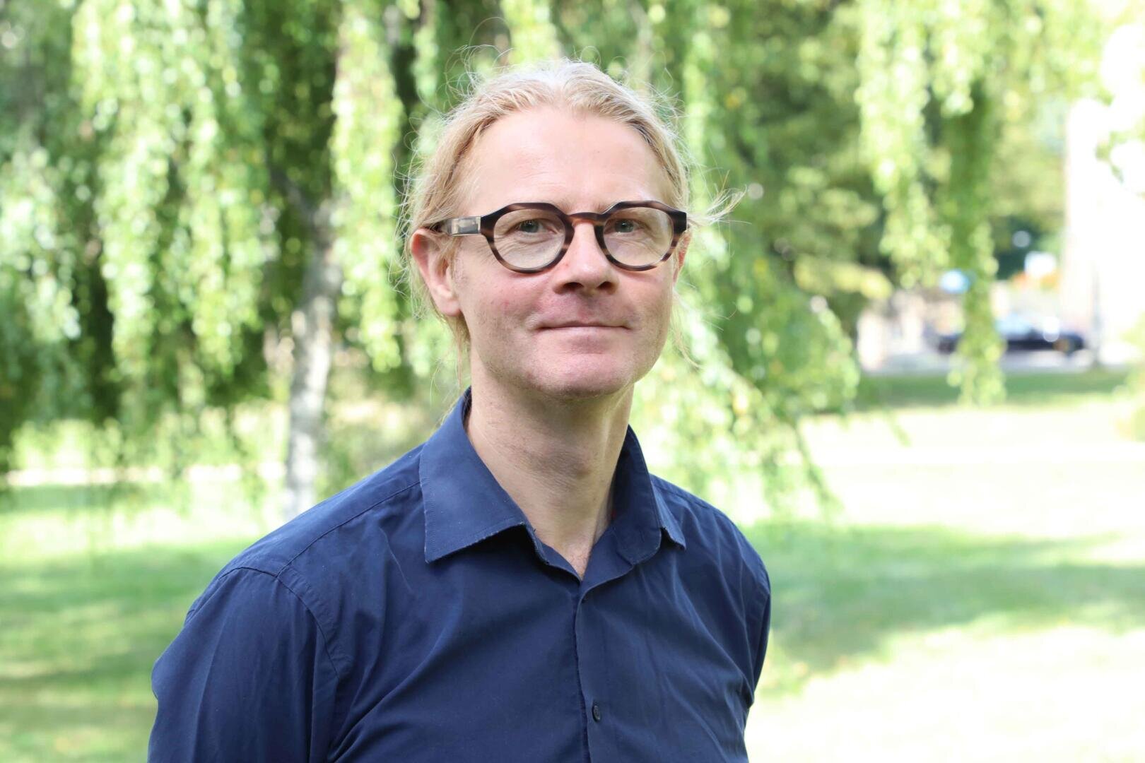 A person with blond hair and glasses, wearing a navy blue shirt, stands outside in a park with green trees and grass in the background.