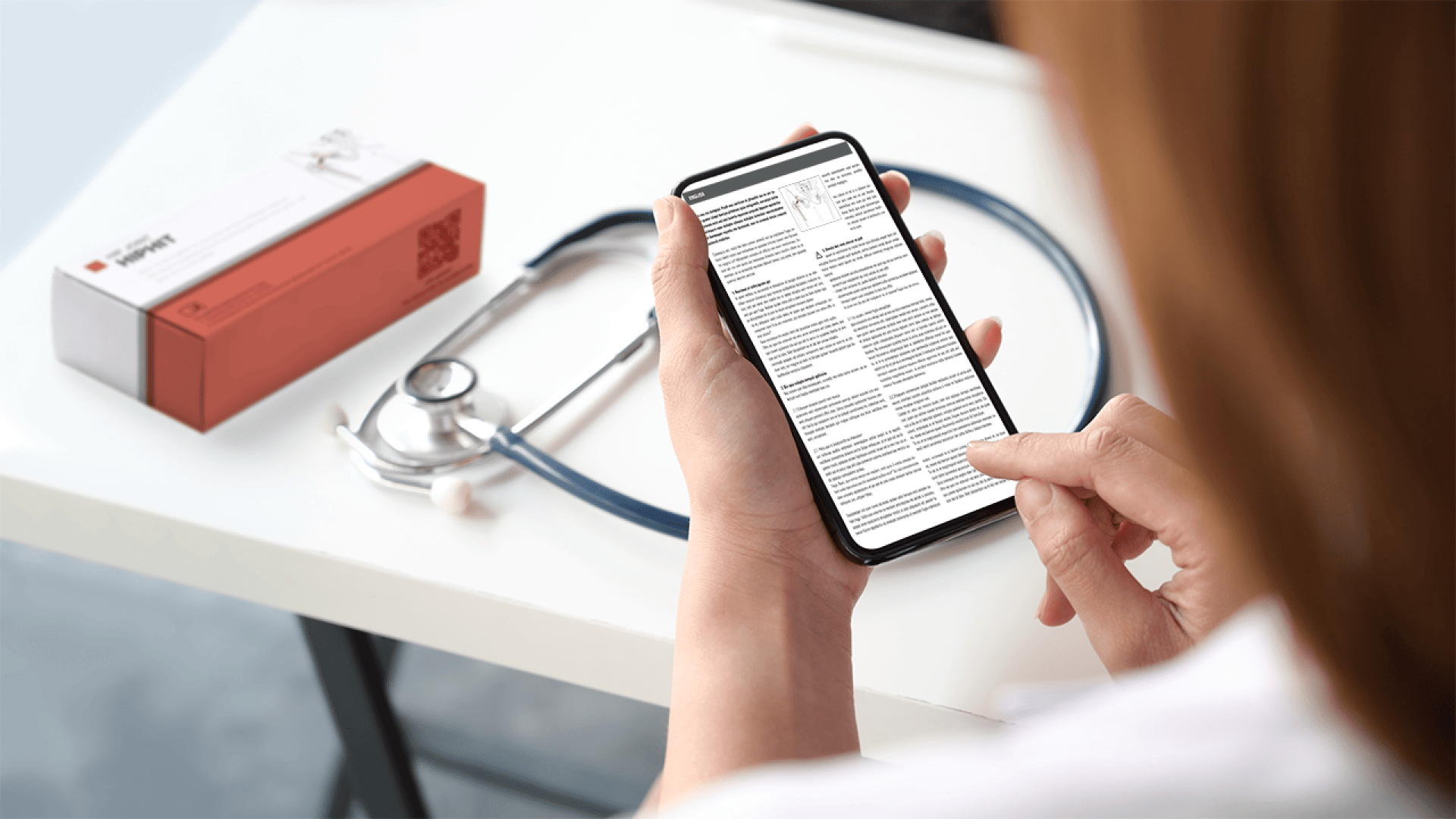 Eine Person in einem weißen Kittel liest mit einem Smartphone Informationen ab. Auf dem Tisch in der Nähe befinden sich ein Stethoskop und eine Schachtel, möglicherweise mit Medikamenten oder medizinischem Zubehör. Die Szene deutet auf ein medizinisches oder gesundheitliches Umfeld hin.