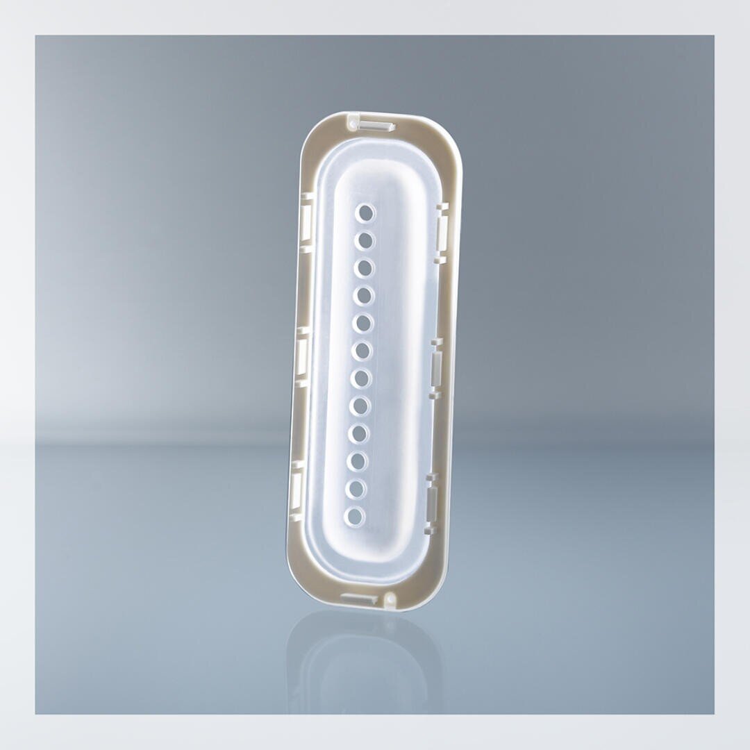 A beige and white rectangular plastic object with rounded edges and two rows of small holes down the center, standing upright on a reflective surface against a light gray background.