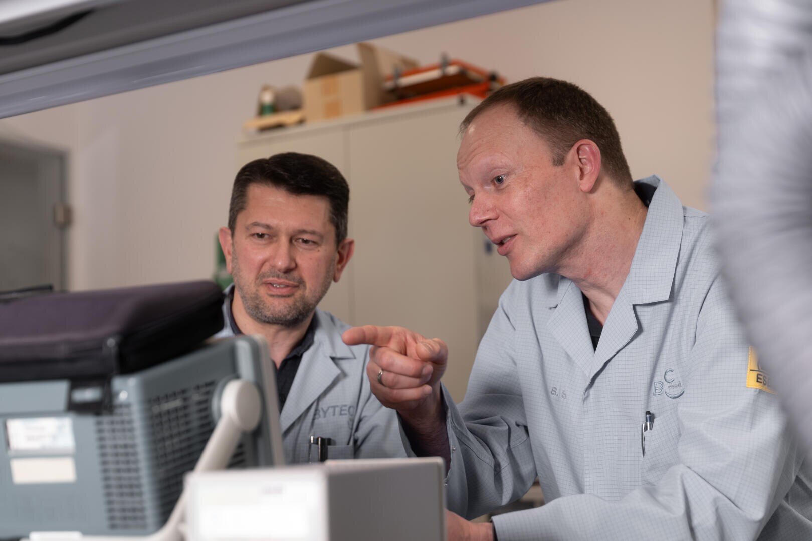 Zwei Männer in Laborkitteln schauen auf einen Computermonitor. Ein Mann zeigt auf den Bildschirm, während er dem anderen, der aufmerksam zuhört, etwas erklärt. Sie scheinen in einem Labor oder einer technischen Umgebung zu arbeiten.