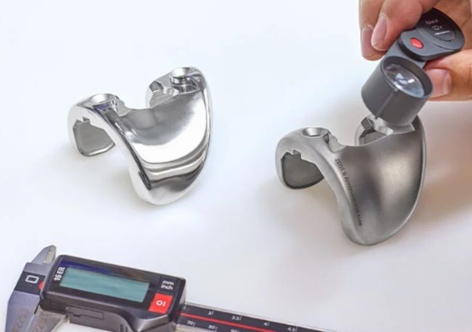 Two shiny metallic knee implants are placed on a white surface. Nearby are a caliper and a magnifying glass held by one hand, indicating that the parts are being checked for quality or precision.