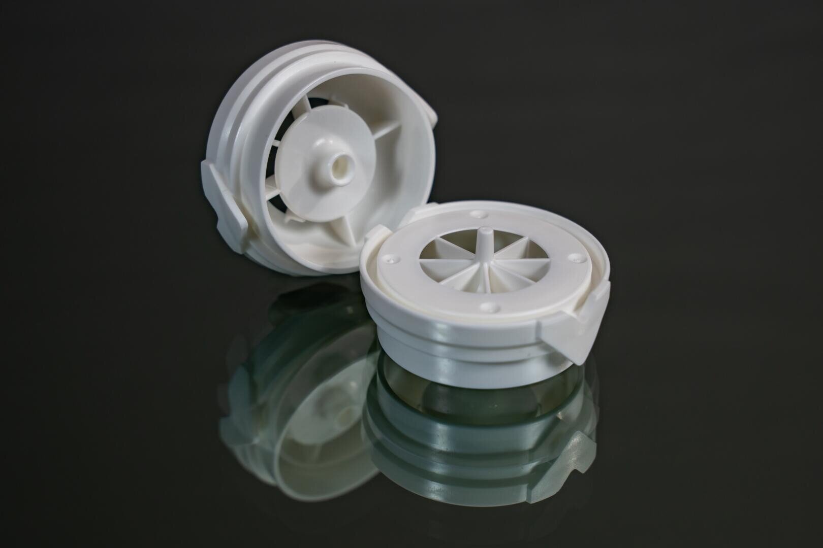 Two white, round plastic objects with radial openings are placed on a reflective dark surface. One is upright, and the other is lying on its side, showing the inner structure.