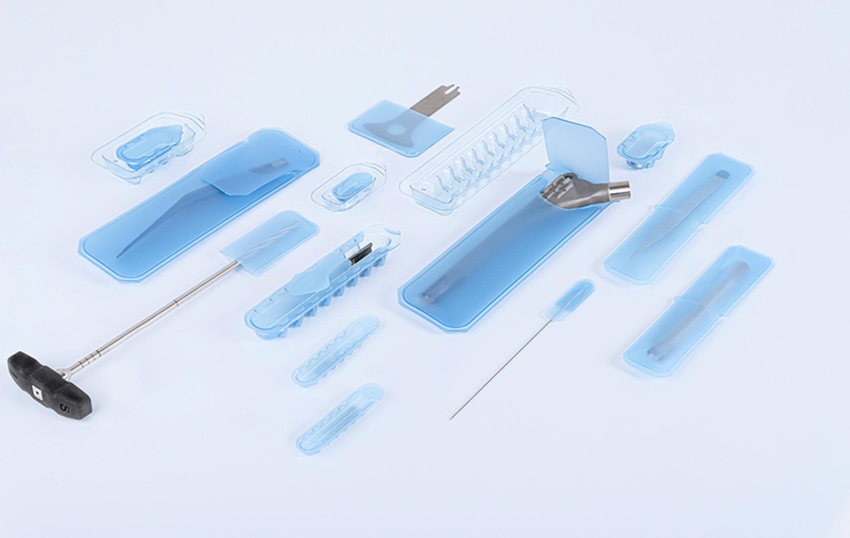 A set of sterilized medical instruments and tools in blue plastic packaging, arranged on a white background. The set contains needles, a handle, a bent metal piece and various small parts.