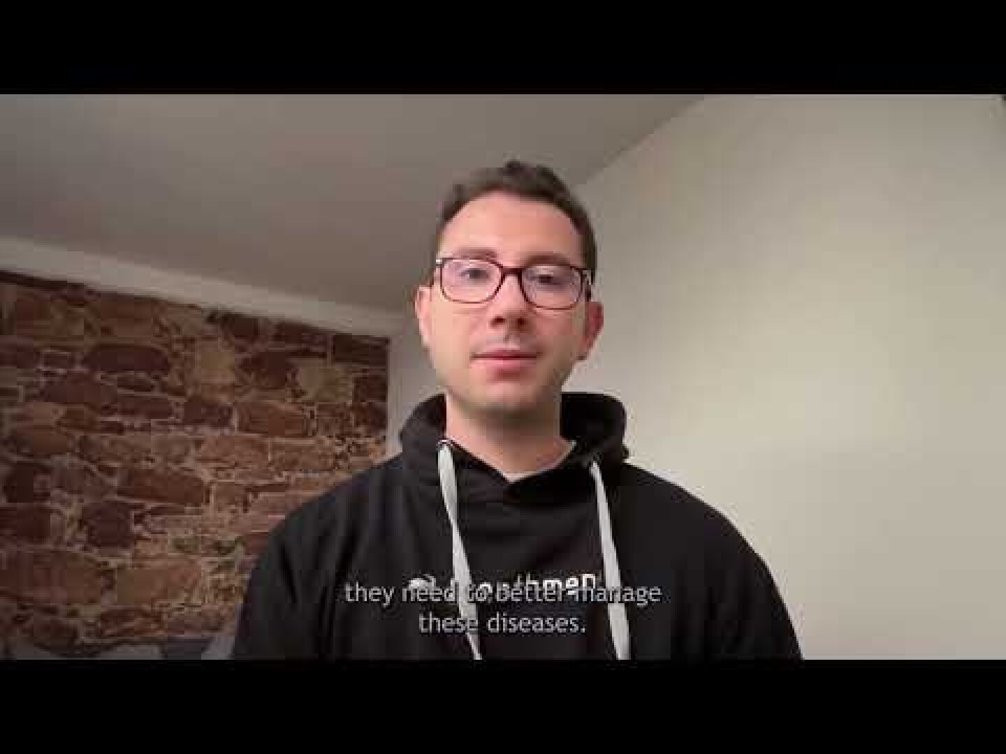 A man wearing glasses and a black hoodie stands indoors against a wall with exposed bricks and white paint. Subtitles at the bottom read, they need to better manage these diseases.