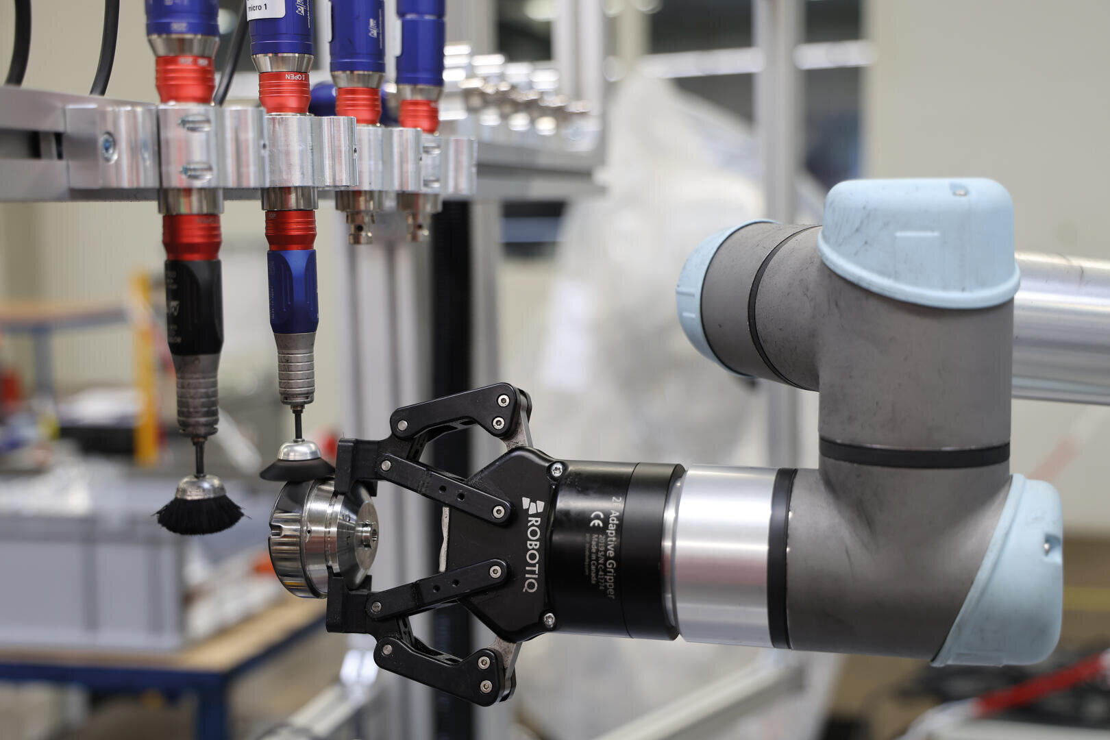 Ein Roboterarm mit einem Robotiq-Greifer hält ein Metallteil in der Nähe einer Reihe von automatisierten Werkzeugen, darunter Bürsten und Bohrer, in einer industriellen Umgebung.