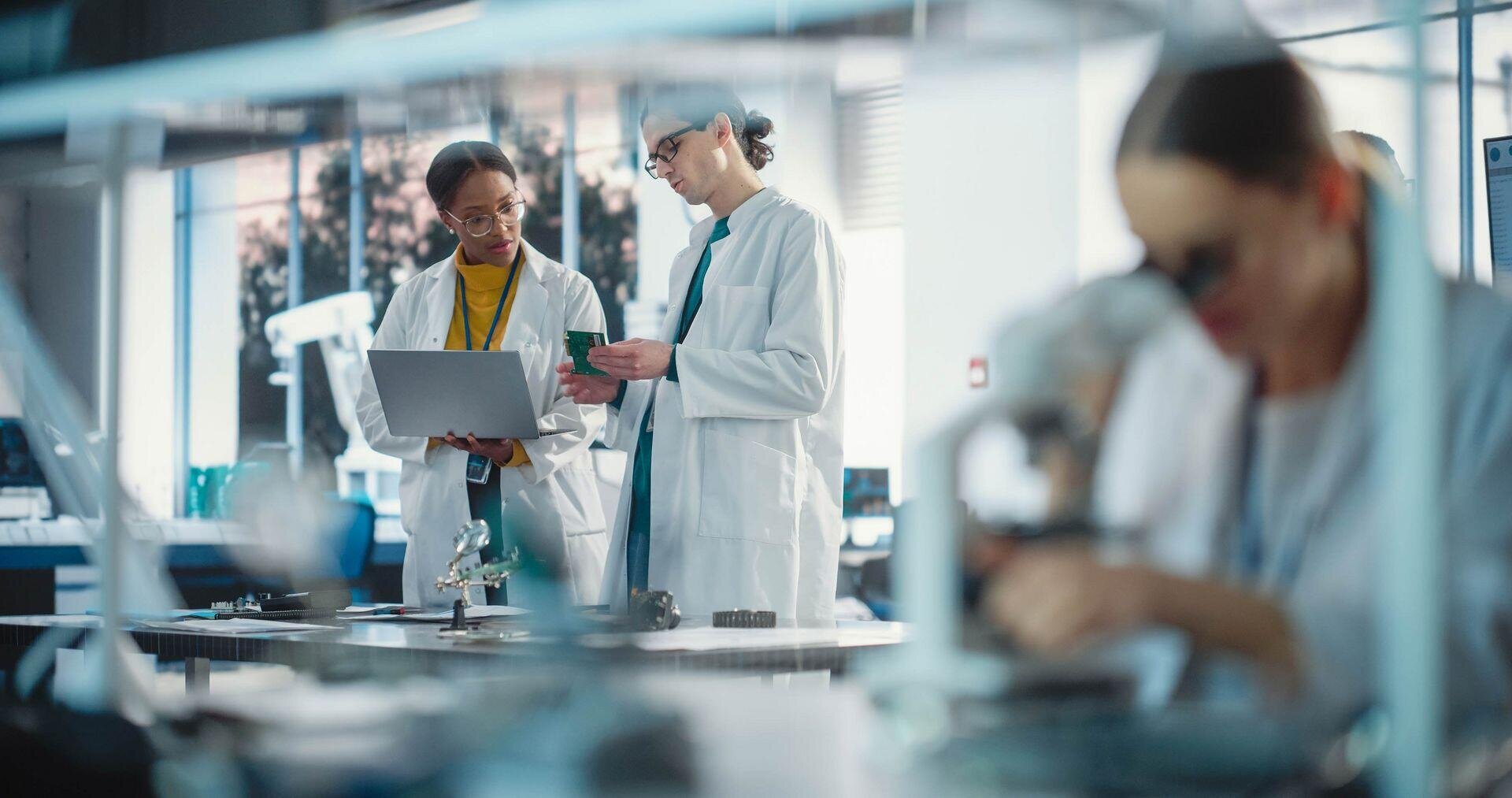 Zwei Wissenschaftler in Laborkitteln diskutieren Daten auf einem Laptop, während eine andere Person im Vordergrund ein Mikroskop benutzt. Auf den Tischen sind Laborgeräte zu sehen, die eine geschäftige, moderne Forschungsumgebung schaffen.