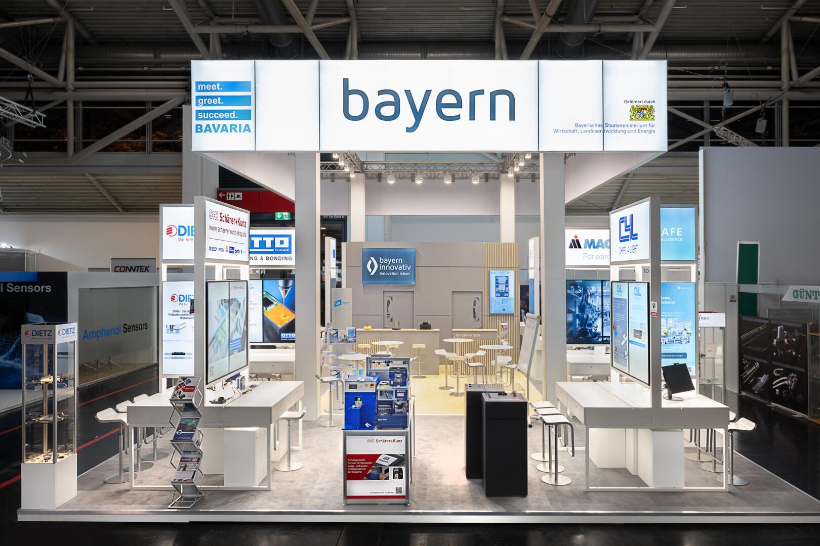 Ein hell erleuchteter Messestand mit einem großen bayern-Schild über dem Kopf, Bildschirmen, Theken, Hockern, Broschüren und Bannern, die Bayerns Technologie- und Innovationsmarken präsentieren.