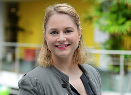 A woman with shoulder-length blonde hair, wearing a gray blazer and a black top, smiles at the camera in a brightly lit room with green plants and a yellow background.