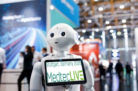 A humanoid robot holds a screen with the words Stuttgart, Germany 2026 MedtecLIVE on a brightly lit trade fair, with blurry people and stands in the background.