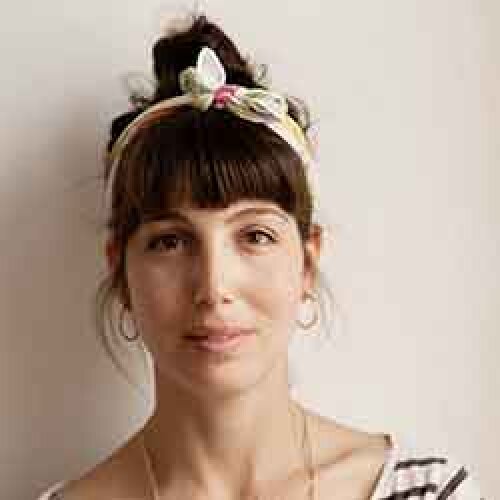 A woman with dark hair in a bun, wearing a flowered headband and hoop earrings, looks neutrally into the camera. She wears a striped shirt and a necklace and stands in front of a plain, light-colored background.