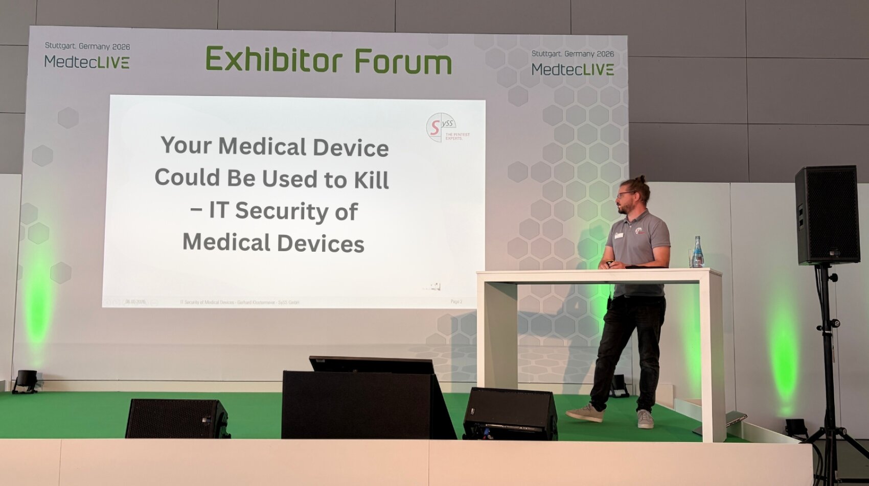 Auf der Bühne des Ausstellerforums der MedtecLIVE 2023 steht ein Redner neben einer Leinwand, auf der eine Präsentation mit dem Titel „Ihr Medizinprodukt könnte zum Töten verwendet werden – IT-Sicherheit von Medizinprodukten“ zu sehen ist.